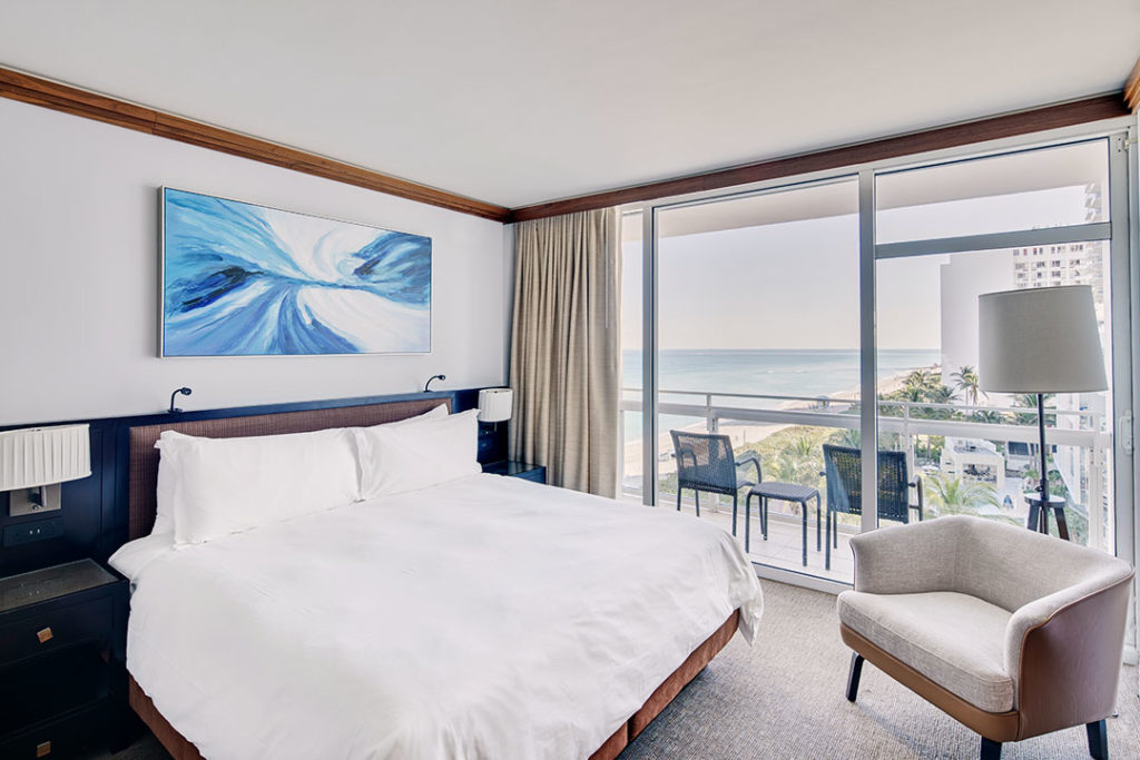 A hotel room with a bed, chair, and balcony overlooking the ocean.