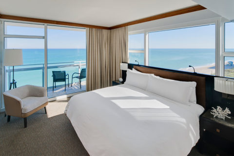 an interior shot of a king bedroom in a suite. The king bed is donned in white linens and is flanked by two black bedside tables. There is a view of the ocean from the balcony.