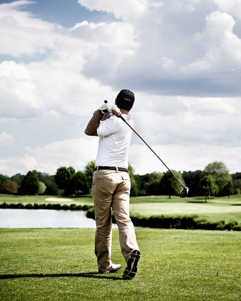 a man with a white collared shirt, khaki pants and back baseball cap follows through on the shot he has just hit playing golf. He looks out onto the expansive green. In the background are sand dunes, a pond and trees