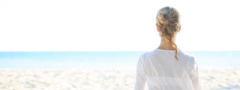 a woman in a white tunic sits cross legged on the white sand. Her hands are resting on her thighs with her palms facing up in the receiving position. She looks out to the ocean