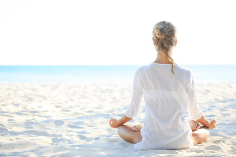 a woman in a white tunic sits cross legged on the white sand. Her hands are resting on her thighs with her palms facing up in the receiving position. She looks out to the ocean