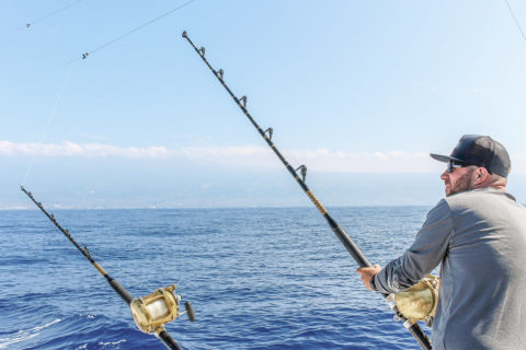 a man faces away from the camera so only his profile shows. He is wearing a gray long sleeved shirt, baseball cap and sunglasses. He is holding a large fishing rod pointed towards the ocean