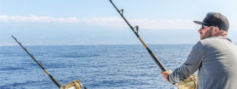 a man faces away from the camera so only his profile shows. He is wearing a gray long sleeved shirt, baseball cap and sunglasses. He is holding a large fishing rod pointed towards the ocean