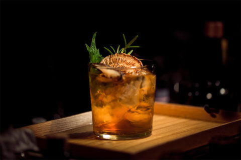 a detail shot of a craft cocktail. The drink is a deep amber color with dried fruit and greens as a garnish. It is in a simple glass on top of a wood platter