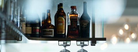 detail shot of a bar shelf. There are bottles of liquor lining the shelf. Below the shelf, wine gasses hang by their stems.
