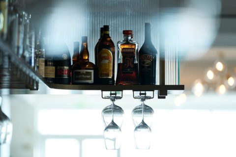 detail shot of a bar shelf. There are bottles of liquor lining the shelf. Below the shelf, wine gasses hang by their stems.