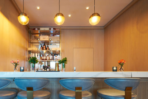 image of the wine bar. Blue and gold chairs line to marble counter top. There are candles and vases with flowers lining the bar. The wall is light colored wood and there are gold shelves with bottle of liquor on them