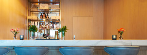 image of the wine bar. Blue and gold chairs line to marble counter top. There are candles and vases with flowers lining the bar. The wall is light colored wood and there are gold shelves with bottle of liquor on them