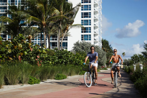 a man and a woman ride carillon branded beach cruiser bikes down a path. The path is lined with greenery on either side. The resort is in the background