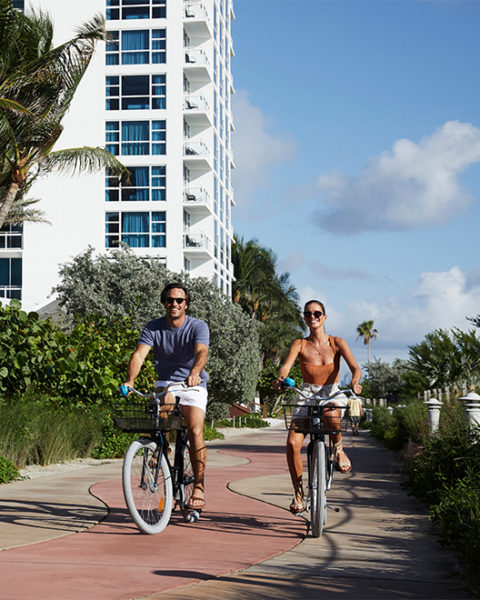 a man and a woman ride carillon branded beach cruiser bikes down a path. The path is lined with greenery on either side. The resort is in the background