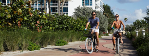 a man and a woman ride carillon branded beach cruiser bikes down a path. The path is lined with greenery on either side. The resort is in the background