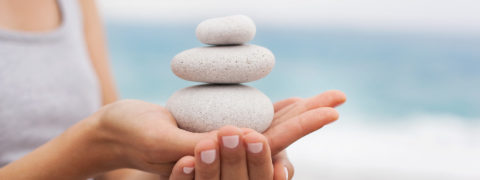 a close up of a woman in a gray tank top's hands. They are placed one ontop of the other. Placed in her palm is a stack of three smooth gray stones. The ocean is visible in the background