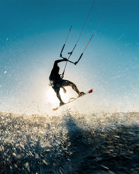 a man on a kite board flies through the air. He grasps on to the bar attached to the kite with one hand. The camera is sprayed with water from the waves he is surfing.