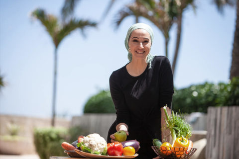 a woman wearing a light green headscarf leans on a table with fruit and vegetable baskets on it. She holds a green apple in her hand. She is smiling at the camera