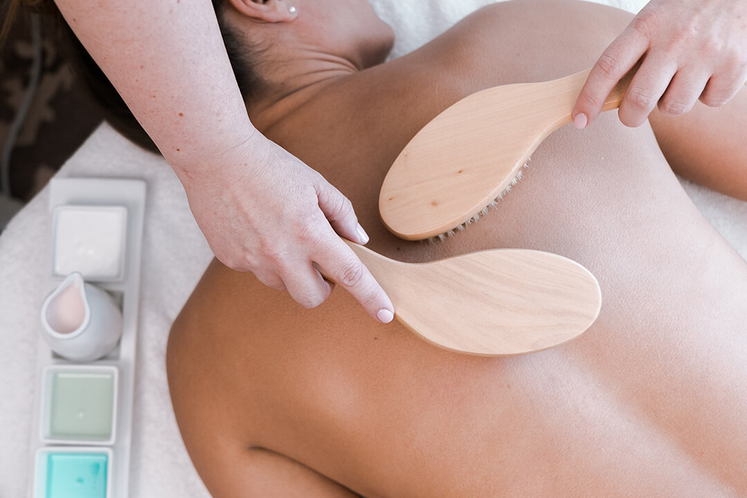 A therapist's hands hold two wooden paddle brushes on a client's bare back.