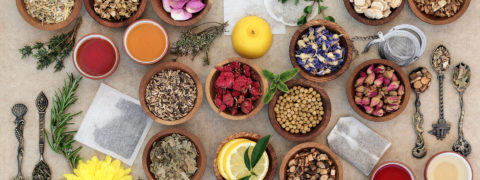 an aerial shot of a beige colored table with a cluster of wooden bowls resting on it. Inside the bowls are different seeds, herbs and floral blossoms. Around the bowls are different green leaves and intricate spoons