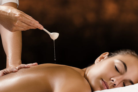 a woman lies face down on a massage bed. Her face is tilted towards the camera. Her eyes are closed and she has a relaxed expression. Above her, a pair of hands is holding a spoon with hot wax slowly dripping from it onto her bare back.