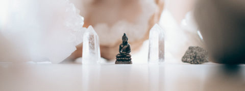 detail shot of a small statue of the Buddah. The statue is a dark color. On either side of the statue are clear crystals.