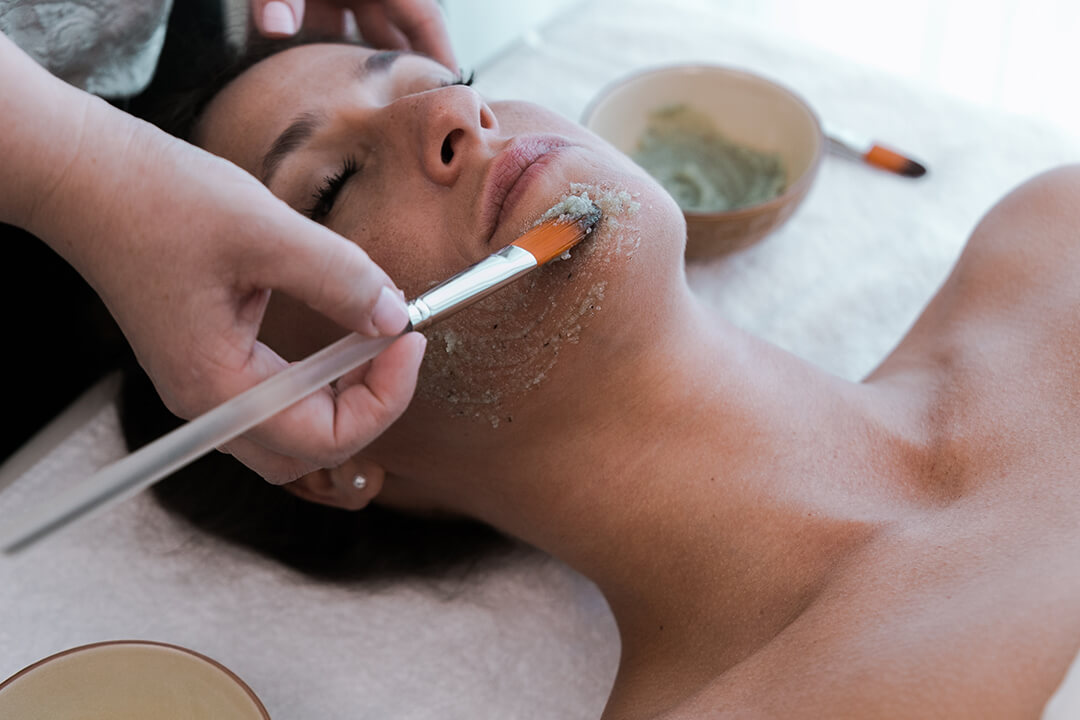 A therapist's hand brushes a pale green mask onto a client's cheek and jaw.