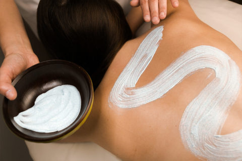 a woman is lying face down on a massage table. Her brown hair is swept to one side leaving her back bare. A pair of hand is applying a light colored clay substance to her back in a wave pattern