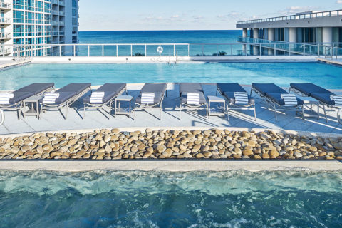 A exterior shot of the Atlantic Spa. In the foreground there is water that is bubbly due to jets in the pool. Looking out from the hot tub, there is a line of gray lounge chairs with striped towels on each of them. They overlook a lap pool, the ocean and the resort buildings on either side