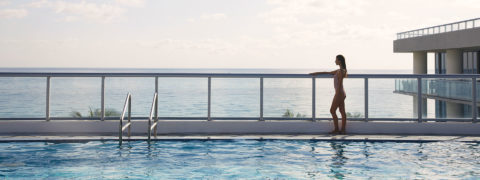 a woman in a bathing suit stands looking out over a balcony. There view is of the ocean and the resort grounds. In the foreground is a long swimming pool