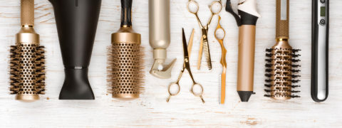 a detail shot of sleek hair care tools. From left to right there is a black hairdryer, a gold barrel brush, a gold spray bottle, two gold scissors, a gold curling iron, a black and gold brush and and black and silver hair straightener. They are all resting on a white wood table.