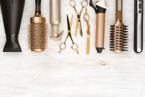 a detail shot of sleek hair care tools. From left to right there is a black hairdryer, a gold barrel brush, a gold spray bottle, two gold scissors, a gold curling iron, a black and gold brush and and black and silver hair straightener. They are all resting on a white wood table.