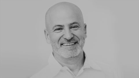 a man in a white button down looks into the camera with a bright smile. The photo is in black and white.