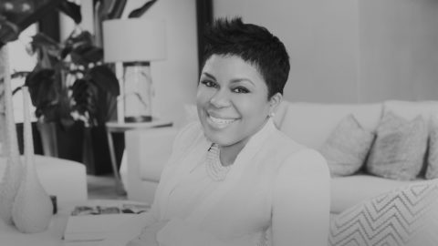 a woman with short hair wearing a white jacket and pearls sits on a couch with patterned throw pillows. She is invitingly smiling at the camera. The photo is in black and white.