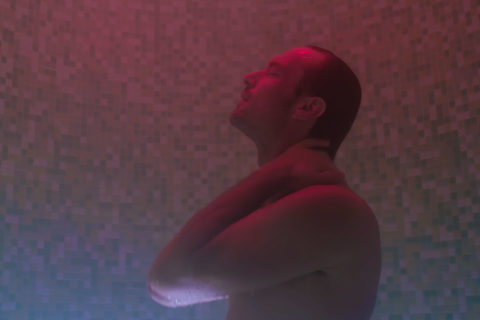 a man stands in a tiled room. He is slick from the water pouring down on him. There is infrared lighting that shines red off of the man's skin and makes the tiles in the background look luminescent.