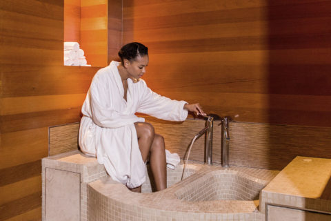 a woman soaks her feet in the foot spa. The bath has tan tiles and a silver water spout. The room is dark wood paneled and there is a nook to store folded white towels. The woman has her hair tied back and is dressed in a white plush robe.