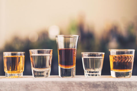 shot glasses of different shapes and designs are lined up on a balcony. The shot glasses are full of dark and clear mezcal and tequila.
