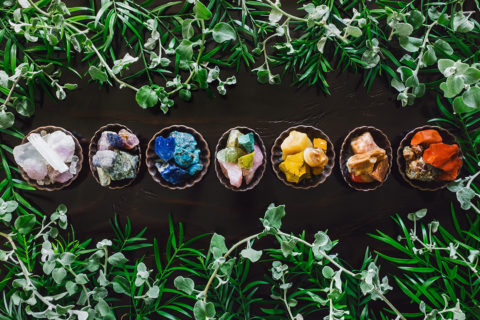 there are seven bowls in a line with different colored mineral rocks piled within them. The minerals are separated by colors such as pink, blue, yellow and orange. The bowls are lined up on a dark wooden table with greenery framing the photo