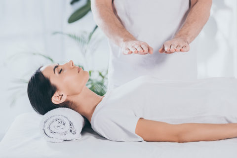 a woman in a white tunic is aying down on a massage table. Her head is propped up on a folded towel and her eyes are closed. There is a man in a white tunic standing over her with his hands hovering over her torso. They are participating in an energy ritual.