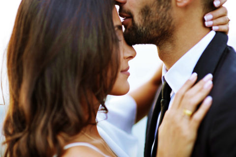 close up shot of a man and woman embracing. The woman is wearing a white dress and the man is wearing a black suit. The woman's hand shows a gold ring on her ring finger.