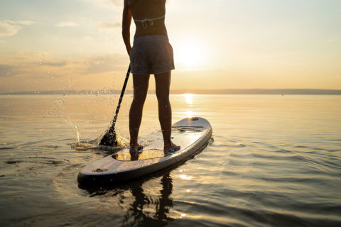 A woman stands on a paddle board in the ocean during sunrise. There is movement in the water as she uses the paddle to move forward. She is wearing a bikini top and cloth shorts