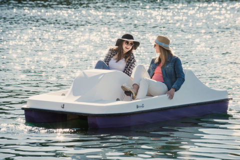 two women are paddling on a blue and white paddle boat in the middle of the water. They are wearing hats and sunglasses and are looking at each other smiling and lauging