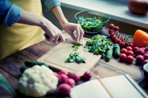 close up shot of a chef with a yellow striped apron on finely chopping herbs on a wooden cutting board. The table surrounding the cutting board is full of colorful, fresh vegetables such as tomatoes, radishes and peppers