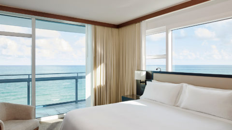 an ocean view suite with a balcony. The bed is on the right side of the room with crisp white linens. There is a black bedside table and a light colored chair in the left corner.
