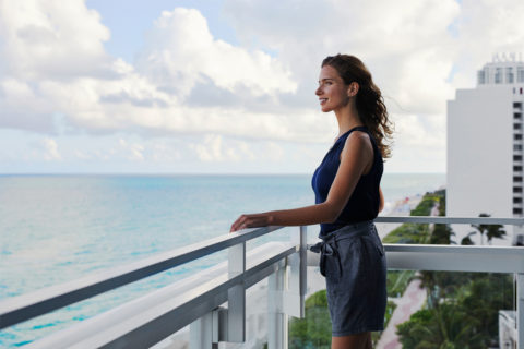 a woman stands on a balcony overlooking the ocean. She is turned so her profile is visible. Her long hair is blowing in the wind and she is smiling. She is wearing loose chambray shorts and a dark blue tank top.