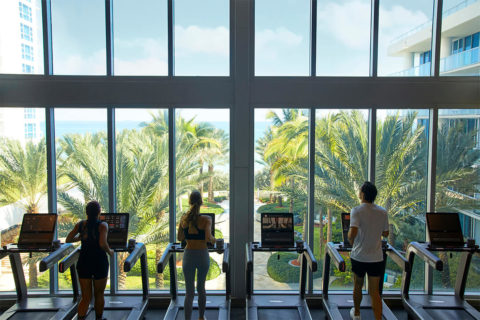 Image of the gym facilities. The shot focuses on the treadmills. There are six treadmills lined against full length windoes with a video of a courtyard that leads to the beach. There are three people in workout outfits running on the treadmills, two woman and one man.