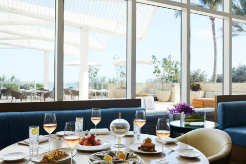 an interior shot of a dining room. There are full length windows that give a view of an outdoor dining area. The seating is plush, blue benches and white leather chairs. The circle table is full with rose glasses at each place setting and multiple dishes in the middle of the table.