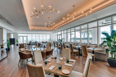 interior shot of a dining room. There are tables lining a gray couch with patterned throw pillows. Each table has blue water cups and wine glasses at each place setting. There are green plants in the background