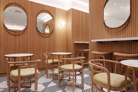 interior shot of a dining room. The floor is black and white tiles and the walls are paneled with light wood. The chairs are light wood with yellow, geometrically patterned cushions. The tables are white and gold circle tops