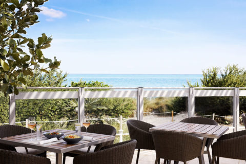 photo of an outdoor dining area. The chairs are woven brown and the tables are a dark wood. At on table there is a glass of rose at each place setting and there are two gray bowls of food in the middle of the table. The outdoor seating area overlooks the sand dunes and the ocean