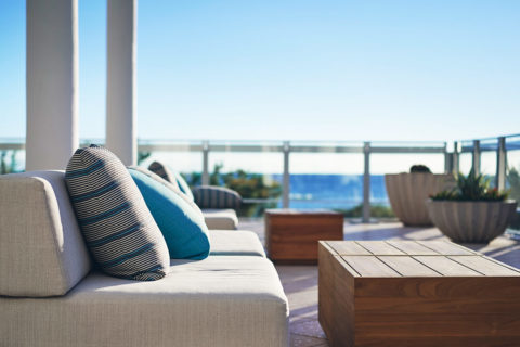 outdoor lounge space with gray cushioned couches accented with blue throw pillows. There are wooden tables in front of each couch and planters with greenery in them. Look over the balcony the ocean is in view.