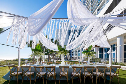a long table setting. there are brown, woo chairs lining the long table. There are three large white vases with green and white flowers flowing from the top. There are tall, crystal candle holders in the middle of the table. The ocean is present in the background. overhead there are white flowers hanging from a canopy