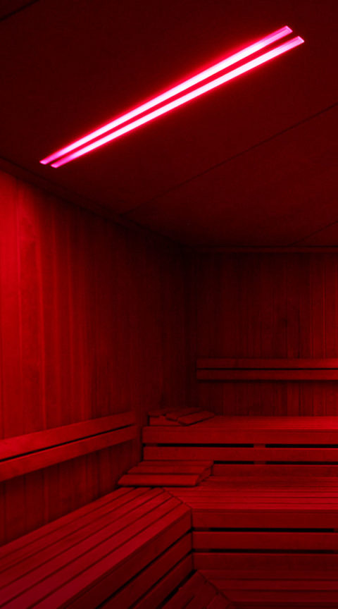 interior image of a thermal therapy room. There are three levels of wooden benches. There is a long infrared light attached to the ceiling that gives off a red glow around the room