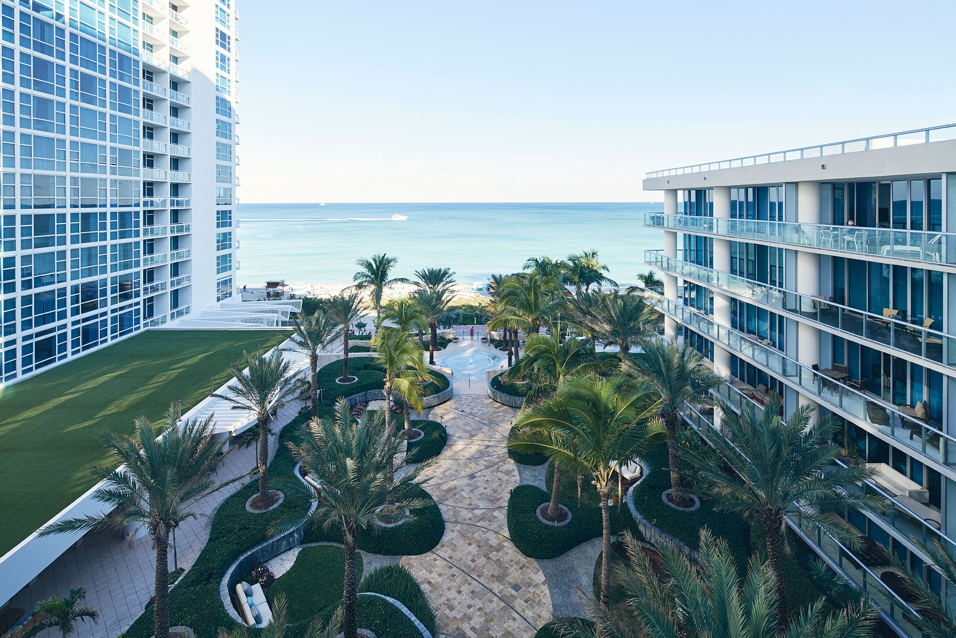 aerial view of the hotel's courtyard. There is a walkway leading to the beach with palm trees and landscaped beds lining the walkway. On either side of the courtyard are rooms with balconies overlooking the it